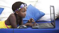 Afrikanisches Mädchen liegt auf dem Bauch vor dem Laptop und schaut auf den Bildschirm.