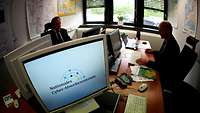 Computermonitor mit Logo des Nationalen Cyber-Abwehrzentrum, im Hintergrund zwei Männer am Computer 