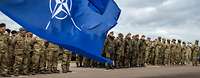 NATO- und EU-Flagge wehen im Wind, im Hintergrund angetretene Soldaten unterschiedlicher Nationen