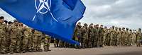 NATO- und EU-Flagge wehen im Wind, im Hintergrund angetretene Soldaten unterschiedlicher Nationen