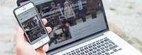 Hand mit Smartphone, im Hintergrund ein Laptop