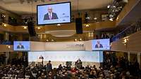 Ein man spricht in einem großen Saal am Rednerpult.