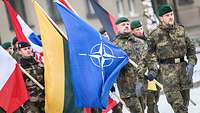 Soldaten unterschiedlicher Nationen marschieren mit Flaggen, ein deutscher Soldat trägt die NATO-Flagge