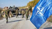 Soldaten auf Patrouille, im Hintergrund gepanzerte Fahrzeuge, im Vordergrund die NATO-Flagge