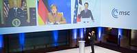 Ischinger steht vor großen Monitoren, auf denen Joe Biden, Angela Merkel und Macron zu sehen ist