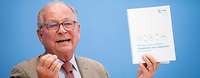 Wolfgang Ischinger sitzt vor einer blauen Wand und hält Bericht hoch