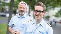Im Vordergund, rechts, ein Soldat im Dienstanzug. Im Hintergrund, links, ebenfalls ein Soldat im Dienstanzug. Beide lächeln. 