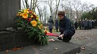 Ministerin Kramp-Karrenbauer hockt mit gesenktem Kopf vor einem Blumenkranz auf dem jüdischen Friedhof.