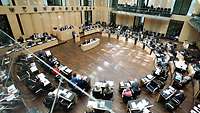 Mehrere Menschen sitzen in einem Saal während einer Versammlung, von oben fotografiert. 