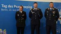 Zwei Soldaten und eine Soldatin stehen in Dienstanzügen vor einer Wand mit der Aufschrift „Tag des Peacekeeping“.