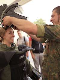 Eine Zivilistin zieht zur Probe den Schutzanzug der Kampfmittelbeseitiger an, ein Soldat setzt ihr den dazugehörigen Helm auf