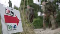 Ein wegweisendes Schild steht am Waldrand, Soldaten laufen daran vorbei.