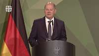 Bundeskanzler Scholz hält eine Rede. Rechts hinter von ihm ist die Flagge der Bundesrepublik Deutschland zu sehen.