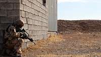 Ein Soldat hockt mit einer Waffe an einer Hausecke, um ihn herum trockene Landschaft.