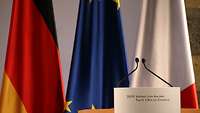 Flaggen von Deutschland, Europa und Frankreich stehen hinter einem Rednerpult.