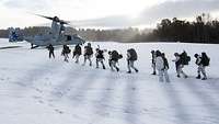 Soldaten marschieren zu einem Kipprotor-Wandelflugzeug, um sie herum Schnee und kahle Bäume, durch die Sonnenlicht bricht