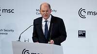 vBundeskanzler Olaf Scholz spricht an einem Podium, man erkennt das Logo der Münchner Sicherheitskonferenz.