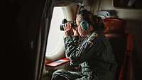 Eine Soldatin beobachtet mit dem Fernglas das Seegebiet aus einem Flugzeug vom Typ P-3C Orion heraus.