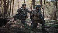 Two soldiers are crouching on the ground of a forest, one is communicating via headset.