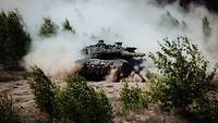 In the terrain between bushes, a camouflaged battle tank Leopard A2 drives, the combat turret is swung to the left.