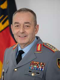 Carsten Breuer schaut in eine Kamera, hinter ihm ist aufgestellt die Flagge Deutschlands.