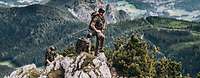 Soldaten stehen auf einer begrünten Bergspitze, weitere Berge und Wälder befinden sich im Hintergrund.