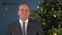 Boris Pistorius spricht in die Kamera, neben ihm steht ein festlich geschmückter Tannenbaum.