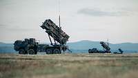 Launcher des Flugabwehrraketensystems Patriot stehen nebeneinander gereiht auf offenem Gelände, drumherum ist Wald erkennbar.