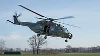Ein Mehrzweckhubschrauber NH-90 MedEvac hebt von einer großen Wiese ab, an der offenen Schiebetür steht ein Soldat.