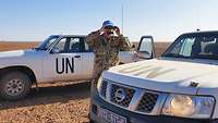 Ein deutscher UN-Soldat schaut zwischen zwei UN-Fahrzeugen durch ein Fernglas.