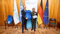 Staatssekretär Hilmer überreicht eine blaue Splitterschutzweste und Helm an einen hochrangigen Soldaten.