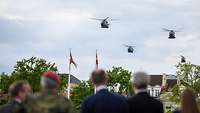 Drei Hubschrauber fliegen über Zuschauer, die unscharf im Vordergrund zu erkennen sind.