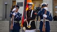 Drei amerikanische Soldaten stehen in einer Ehrenformation nebeneinander, einer hält die deutsche Flagge.