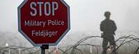 Ein Stopschild steht vor einem mit Nebel überzogenem Gelände, abgeriegelt durch Stacheldraht, dahinter steht ein Soldat.