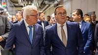 Verteidigungsminister Pistorius geht mit einem Mann im blauen Anzug durch einen großen Raum
