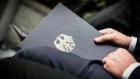 Close-up of a person in a suit, holding a briefcase with an emblem.