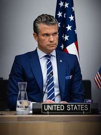 Pete Hegseth sitzt an einem Tisch, davor sein Namenschild sowie amerikanische Flaggen im Hintergrund