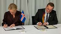Thorgerdur Katrín Gunnarsdóttir und Boris Pistorius sitzen nebeneinander und unterzeichnen ein Dokument.