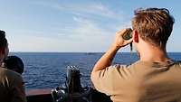 Auf offenem Gewässer beobachtet ein Soldat die türkische Fregatte Gemlik mit einem Fernglas von der Nock aus