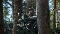 Zwischen Bäumen steht getarnt ein Soldat im Turm einer Panzerhaubitze 2000 und beobachtet die Umgebung.