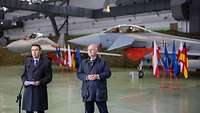 Zwei Männer stehen zusammen vor Mikrofonen. Im Hintergrund stehen zwei Kampfflugzeuge.