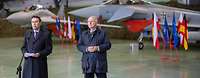 Zwei Männer stehen zusammen vor Mikrofonen. Im Hintergrund stehen zwei Kampfflugzeuge.