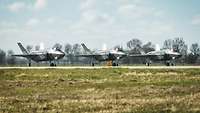 Drei Kampfflugzeuge vom Typ F-35 stehen nebeneinander auf einer Betonfläche.