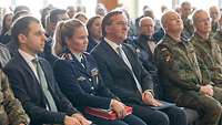 Boris Pistorius sitzt mit vielen Menschen in einem Saal und hört einem Vortrag zu.