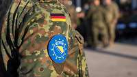 Im Vordergrund ist der Arm eines deutschen Soldaten, mit dem Wappen der Mission EUFOR "Althea".