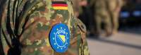 Im Vordergrund ist der Arm eines deutschen Soldaten, mit dem Wappen der Mission EUFOR "Althea".