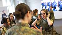 Eine junge Frau setzt sich Kopfhörer auf und probiert Ausrüstung der Bundeswehr an. 