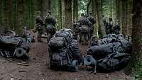 Rucksäcke von Soldaten liegen und stehen auf dem Waldboden. Im Hintergrund sind Soldaten zu sehen.