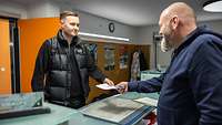 Ein junger Mann bekommt von einem Mann einen Zettel übergeben.