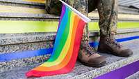 Eine Regenbogenfahne liegt auf einer Treppe, dahinter sind die Beine einer Soldatin zu sehen.